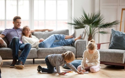 Two children draw on the floor with colored pencils, while two adults relax on a sofa in a bright, modern living room with large windows and plants.