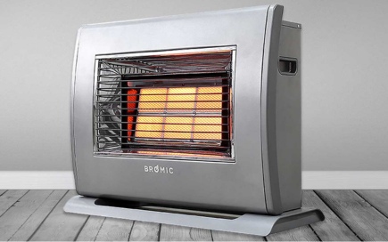 A portable gas heater emits a warm, orange glow through its grated front, positioned on a wooden floor against a plain grey wall. Text: 