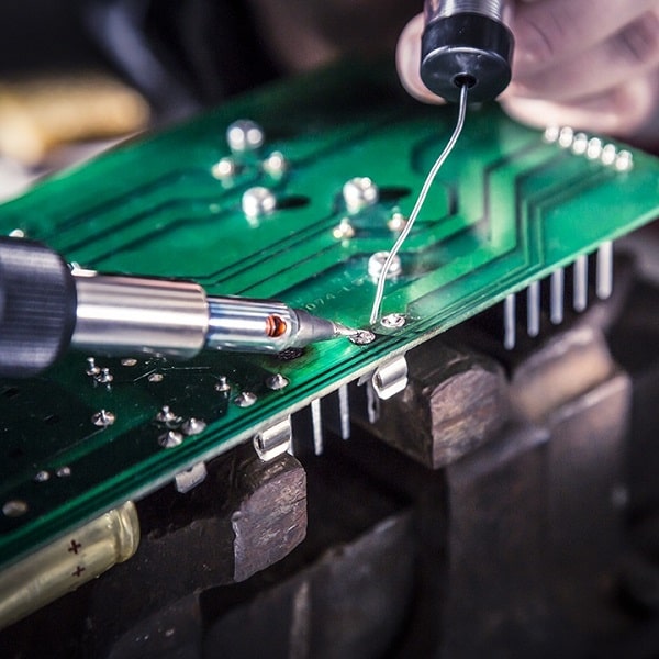 A green circuit board is being soldered using a soldering iron and wire in a workshop setting, with components and tools visible in the background.