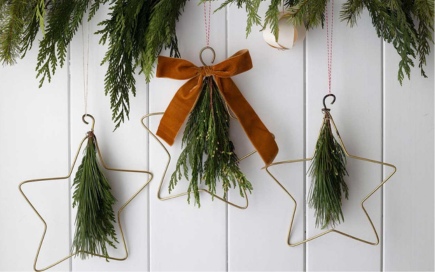 Three star-shaped wire ornaments hang on a white wall, adorned with greenery. The middle ornament features a large brown bow, under a garland of lush greenery.