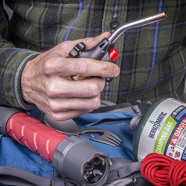 A hand holds a camping lighter above a backpack containing a flashlight, fork-spoon combo, and Bernzomatic camping gas. Nearby, red rope is coiled on the gear.