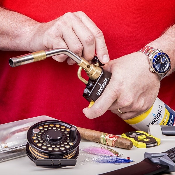 Hands hold a Bernzomatic torch, igniting a flame. Nearby, a cigar, a fishing reel, lures, and a knife lie on a tabletop. The person wears a red shirt and a watch.