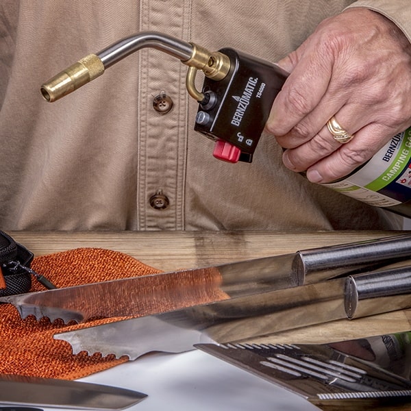 A hand operates a Bernzomatic torch above a serrated knife, on a wooden surface with other tools nearby.