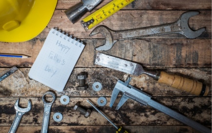 Tools, including wrenches and a caliper, lie scattered on a wooden surface. A yellow hard hat and notepad with "Happy Father's Day" written add context of celebration or gift preparation.