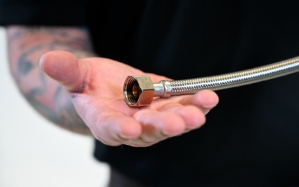 A metal braided hose is held by a tattooed hand, positioned open against a blurred dark background, suggesting inspection or demonstration.