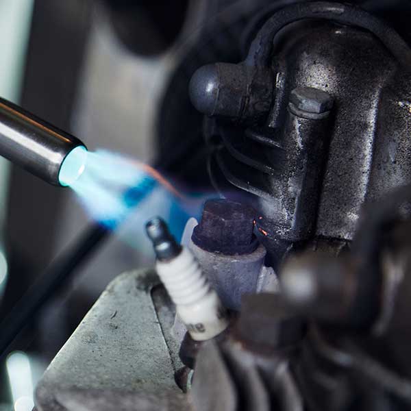 A blowtorch emits a blue flame, heating a metal engine component, with a visible spark plug nearby, within a workshop setting.