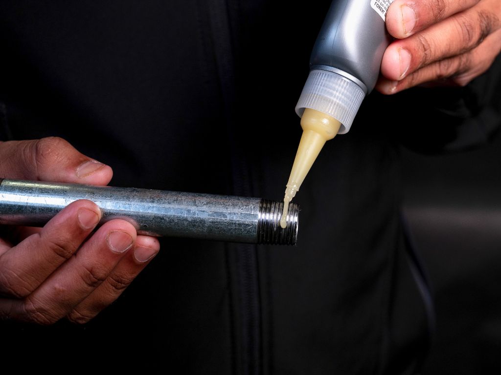 Hands hold a metal pipe while applying liquid from a squeeze bottle. The person wears a dark jacket, and the environment is dimly lit.