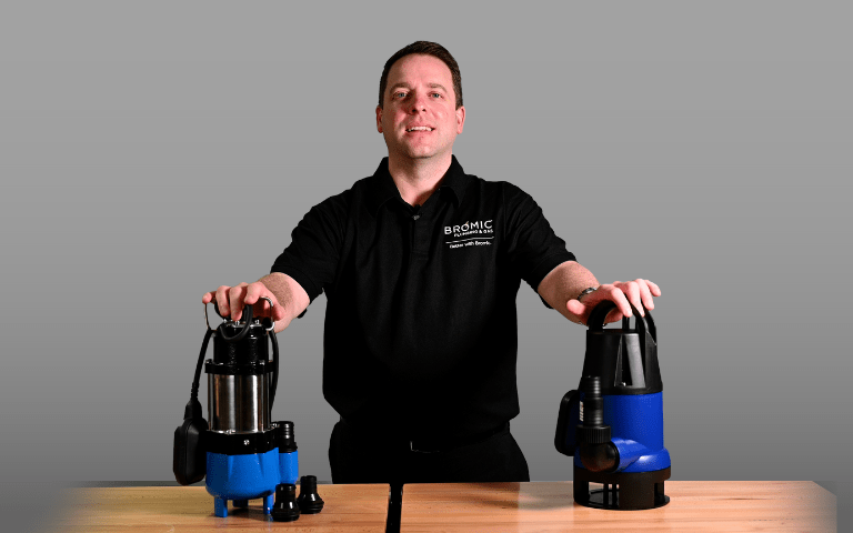 A person stands behind two blue sump pumps on a wooden table, demonstrating the products. The shirt reads, "Bromic: Trade with Confidence" in a plain indoor setting.