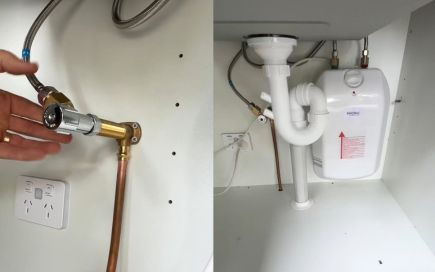 A hand adjusts a brass valve connected to pipes under a sink. Nearby, a white plumbing system and electric water heater are visible inside a cabinet alongside an electrical outlet.