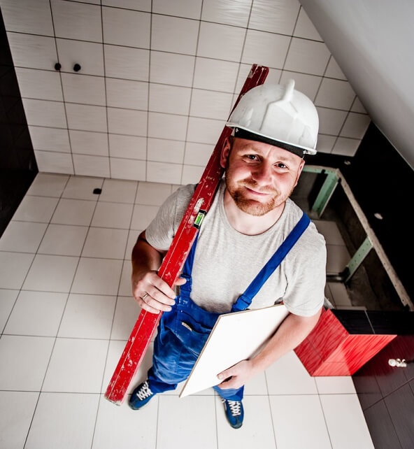 A construction worker, wearing a white helmet and blue overalls, holds a red level and white tile in a room with tiled walls and floor, partially installed fixtures visible.