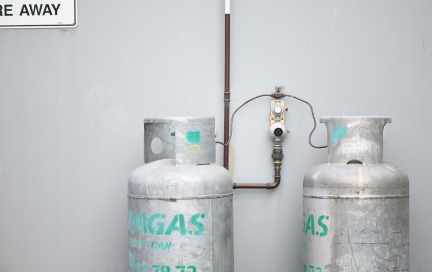 Two large LPG gas cylinders connected to a dual-stage gas regulator mounted on a grey wall.