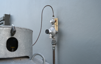 Close-up of a wall-mounted gas regulator system connected to a large outdoor propane tank. The regulator is affixed to a grey exterior wall and linked via piping and a copper sensing line.