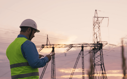 A utility worker wearing a safety helmet and high-visibility vest inspects a device near high-voltage power lines at sunset.
